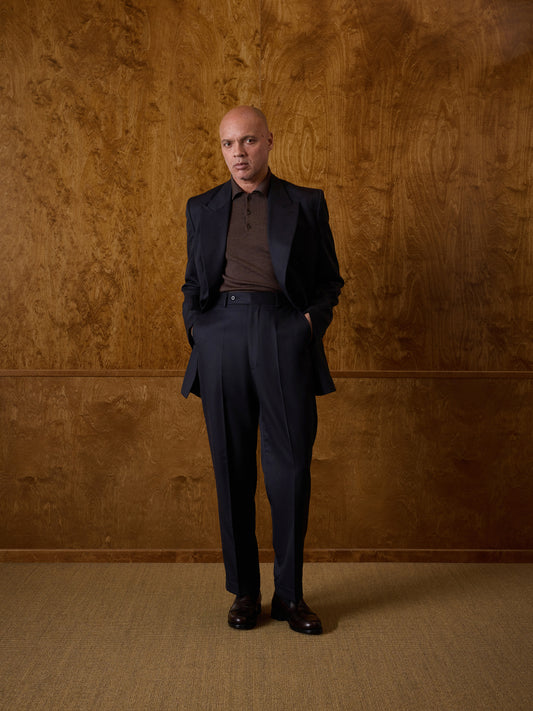 A man wearing a dark navy blue suit, brown merino wool polo, and black shoes stands with his hands in his pockets against a textured brown wood-paneled wall.